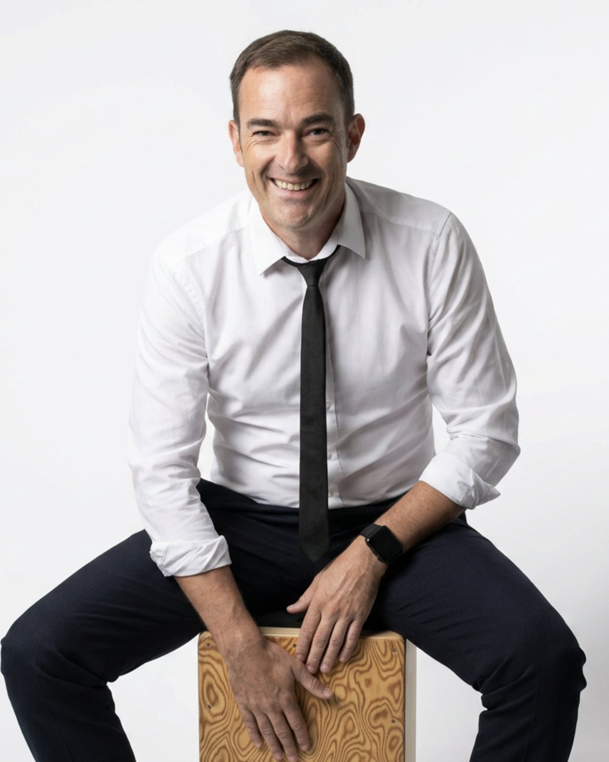 Studio portrait of Matt in a white shirt and black tie, sitting on a cajon
