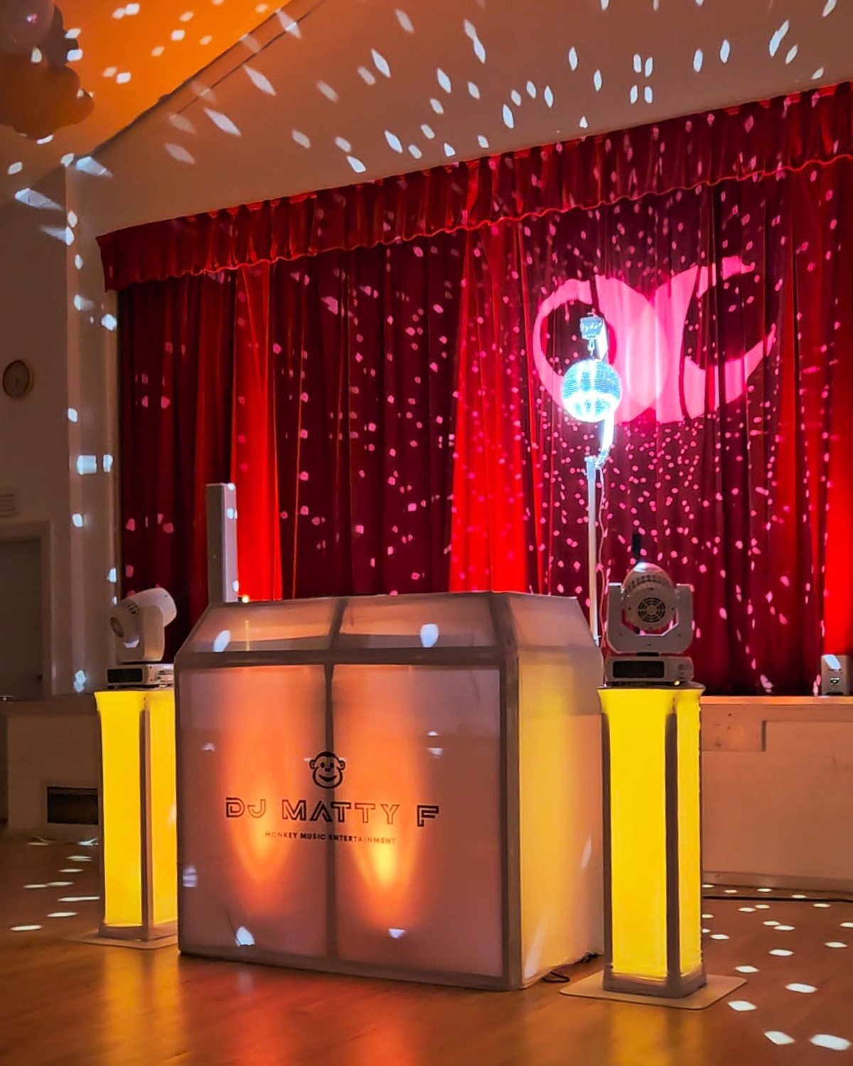 The DJ Matty F booth between yellow uplighters, with a mirror ball and moving heads lighting the room