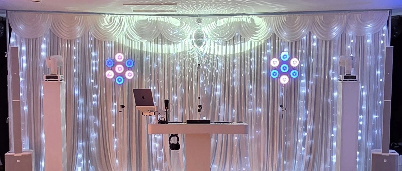 The white DJ booth centred against a starlight backdrop, mirror ball above and uplighters either side